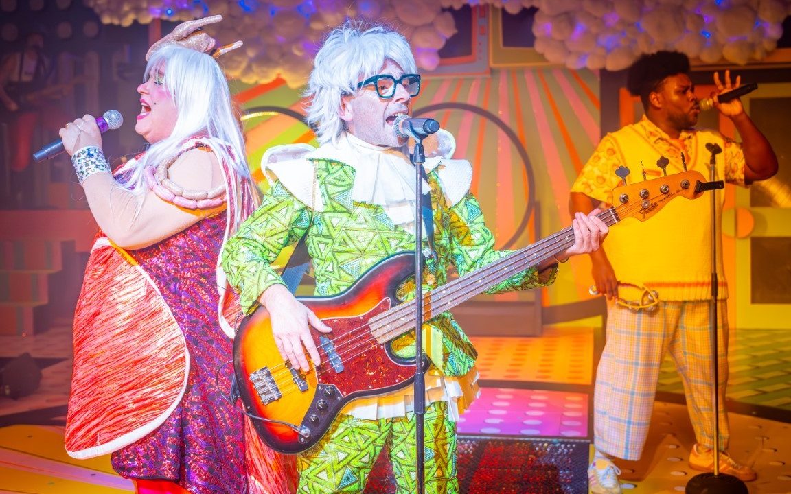 A person wearing an all green outfit with a white jabot playing the electric guitar and singing into a microphone which is on a stand.