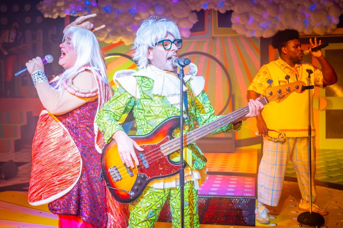 A person wearing an all green outfit with a white jabot playing the electric guitar and singing into a microphone which is on a stand.