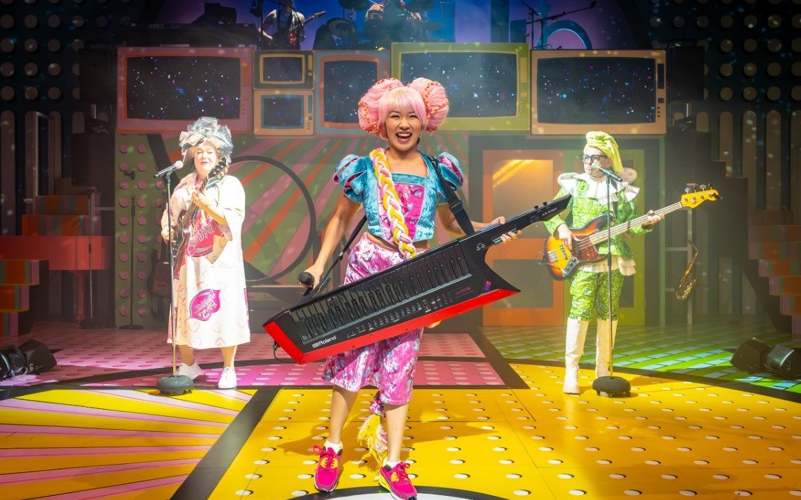 A woman stood with a large electric keyboard around her neck. She has pink hair in two buns, a blue top and pink trousers on. In the background there are two people singing into microphones