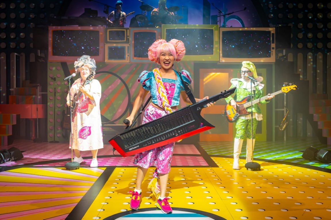 A woman stood with a large electric keyboard around her neck. She has pink hair in two buns, a blue top and pink trousers on. In the background there are two people singing into microphones
