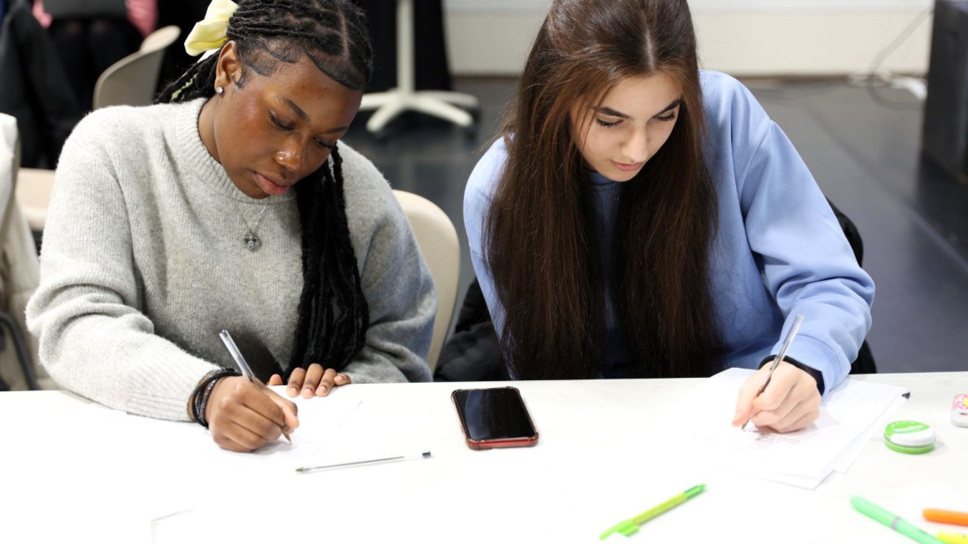 a black and a white teenage girl sat together at a table writing.