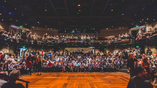 A picture taken from on stage, there is audience filled all around the three sides of the thrust stage