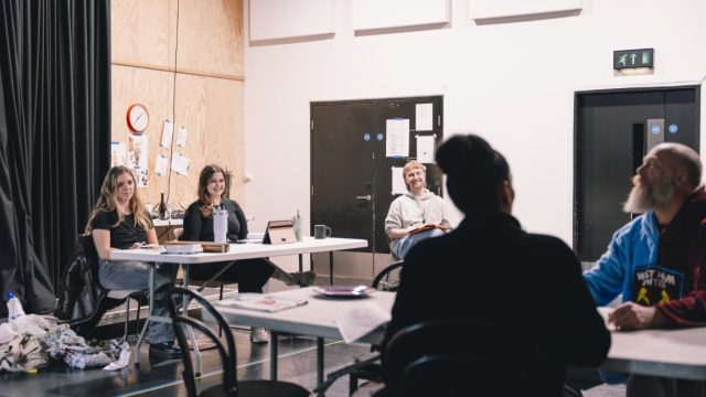 Two young producers sat behind a desk and another sat on a chair separate to the desk in rehearsals. in the foreground there are two people rehearsing for a play.