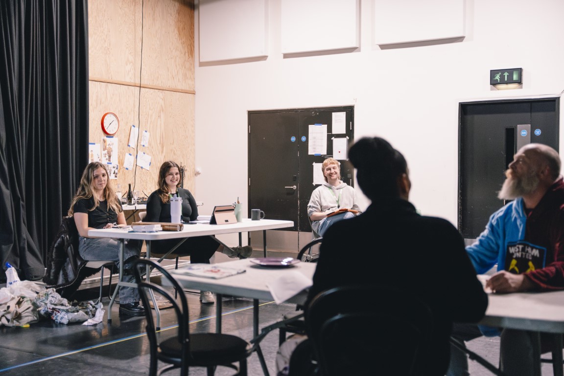 Two young producers sat behind a desk and another sat on a chair separate to the desk in rehearsals. in the foreground there are two people rehearsing for a play.