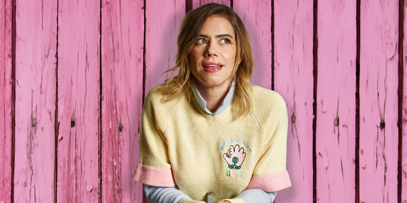 a white woman standing in front of a pink background, she is wearing a yellow jumper and is looking to the right.