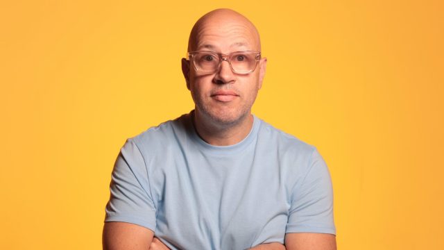 A bald man wearing glasses stands in front of a yellow background with his arms folded