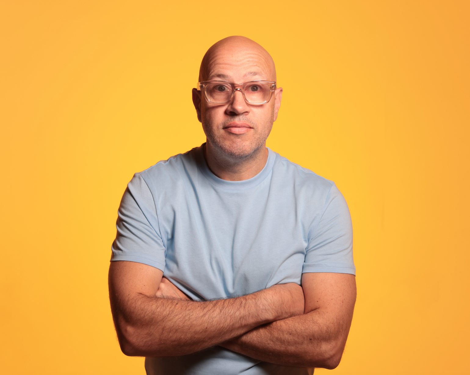 A bald man wearing glasses stands in front of a yellow background with his arms folded