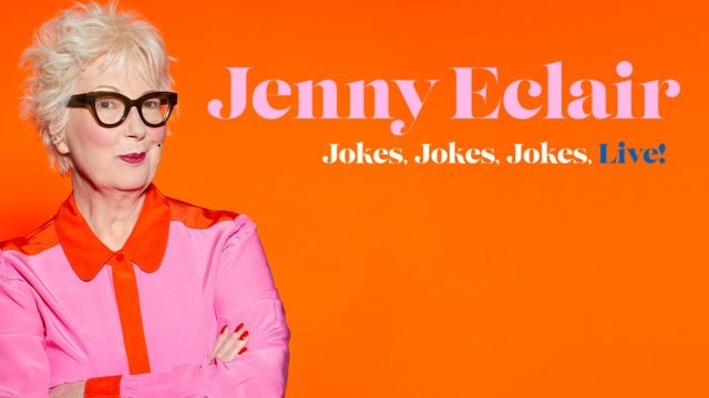 A white woman wearing glasses and a pink and orange shirt, stood against an orange background. The text on screen reads 'Jenny Eclair Jokes Jokes Jokes Live'