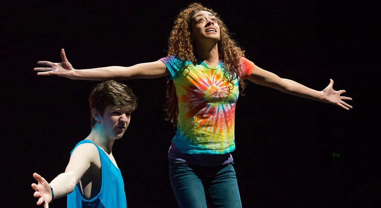 A girl in a tie dye top stood next to a guy with a vest who is kneeling on the floor. They both have their arms to the sides