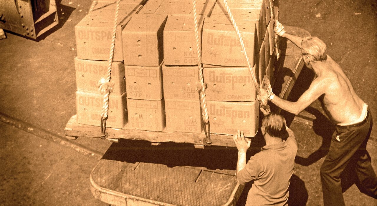 A topless man and a man in shirt lugging boxes on a trolley