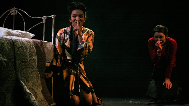 Two people next to a bed crawling on the floor with a finger over their lips