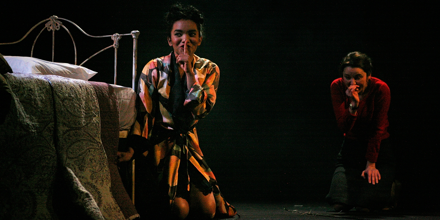 Two people next to a bed crawling on the floor with a finger over their lips