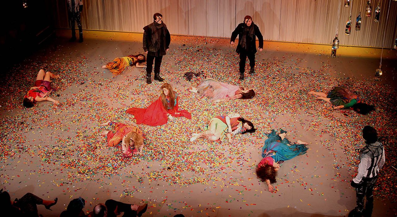 A group of women lying on a confetti coated floor. There are three men stood looking at them