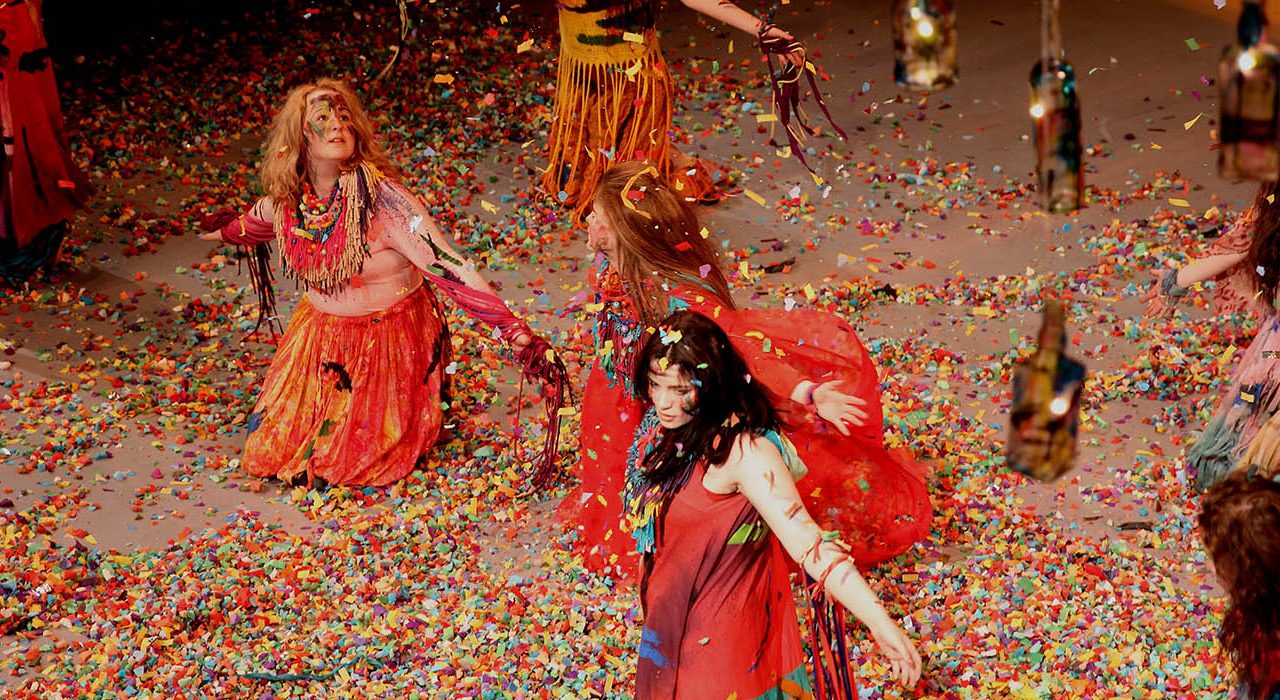 two women wearing floaty orange clothing kneeling in confetti which is falling down on them.