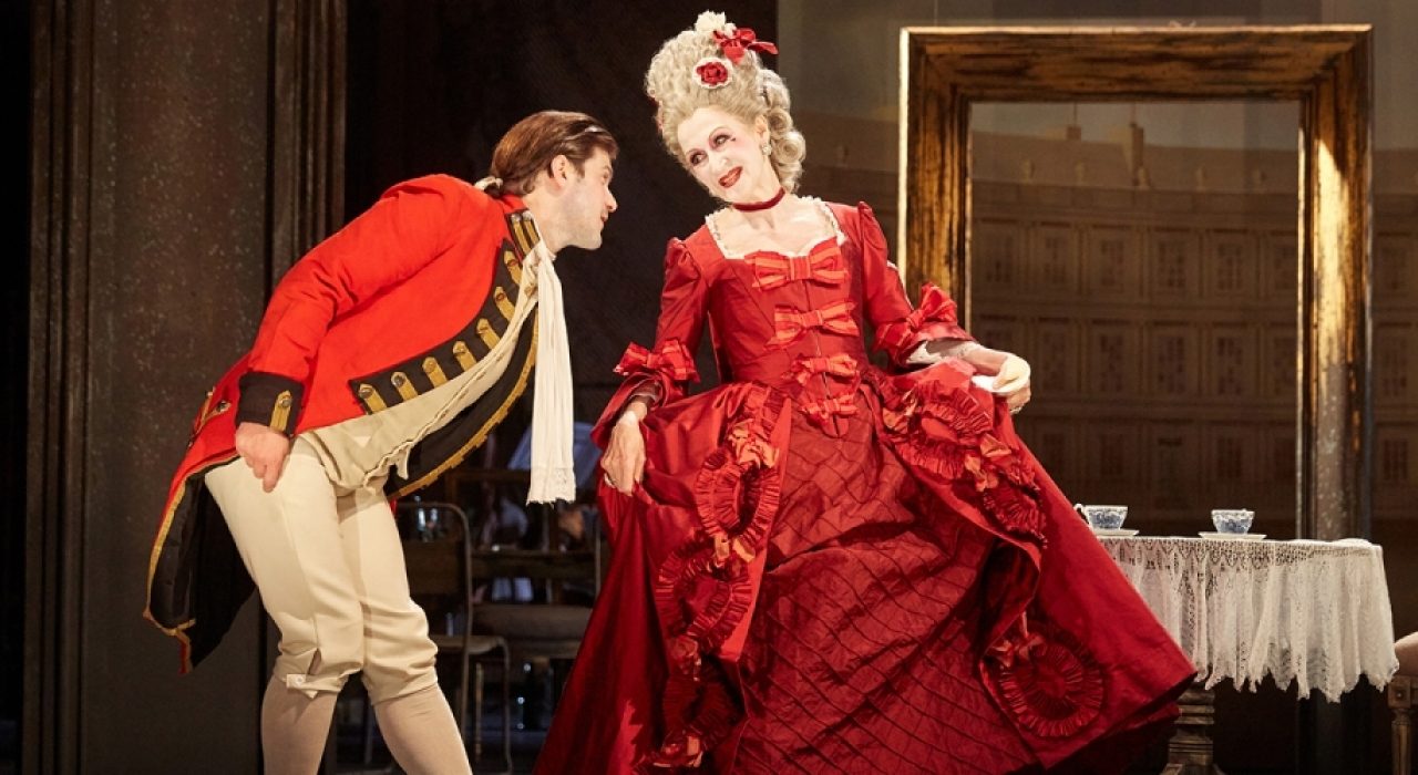 A man in a royal guard outfit bowing at a woman who is wearing a large pink dress and powder wig