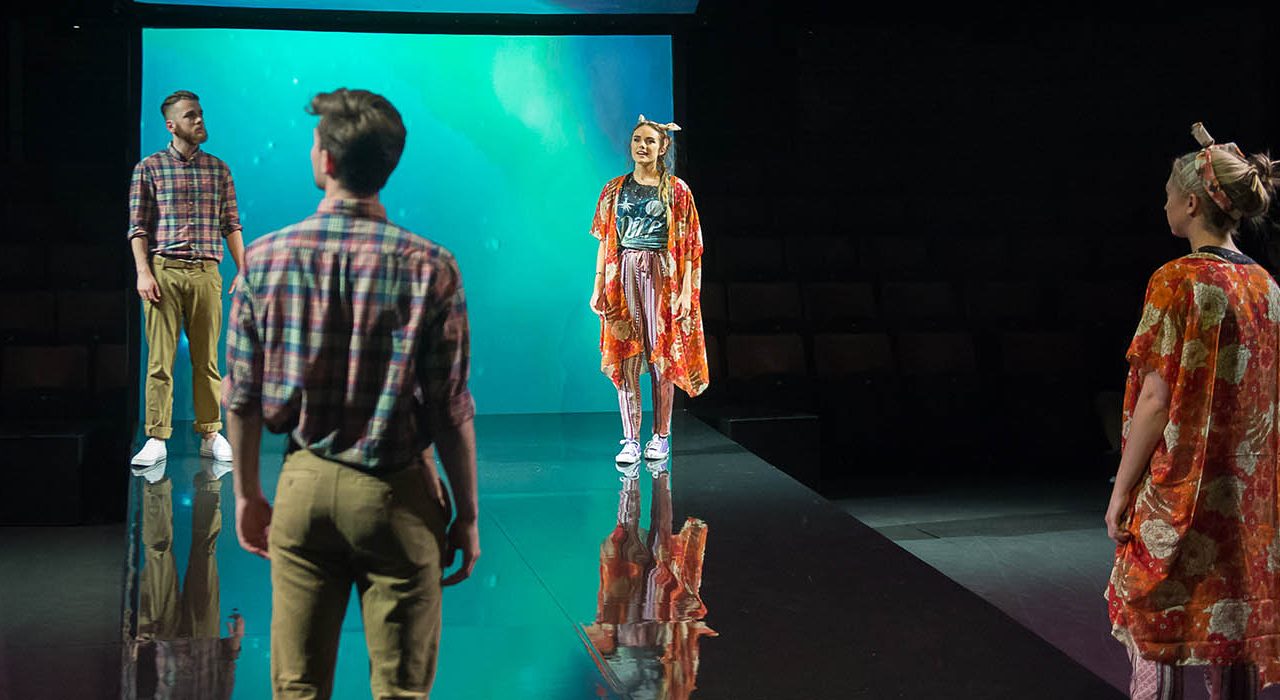 A man in checkered shirt and beige trousers next to a woman with a bow head band and orange jacket. Looking at two people who are clones of them.