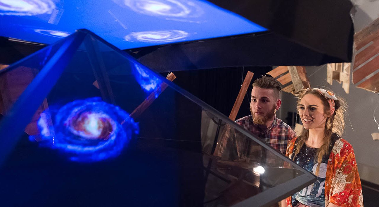 A man and a woman looking at a pyramid that has a projection of the universe in it