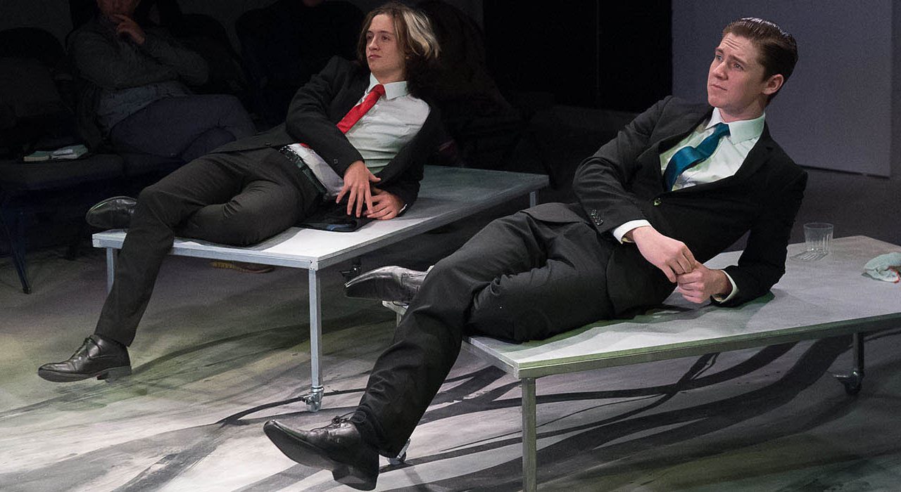 Two men leaning back on tables wearing suits. one has a blue tie and the other has a redo ne