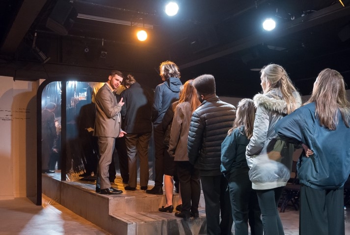 A line of people in coats queuing whilst a man in a suit ushers them in