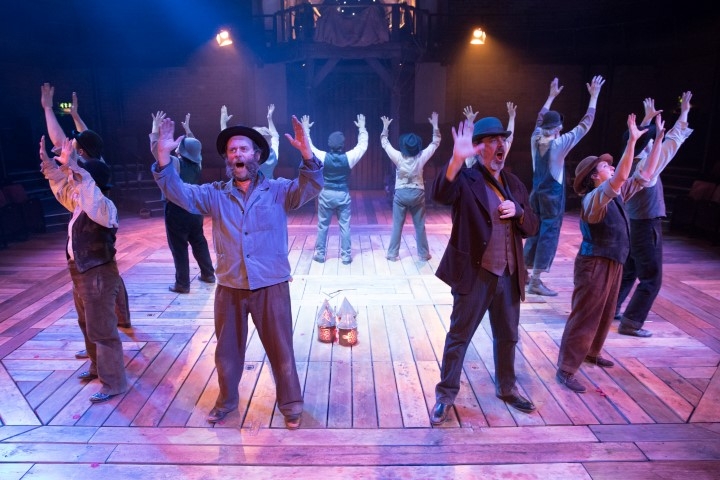 A group singing dressed in shirts waist coats and hats. They are in a circle formation facing outwards with their arms raised.