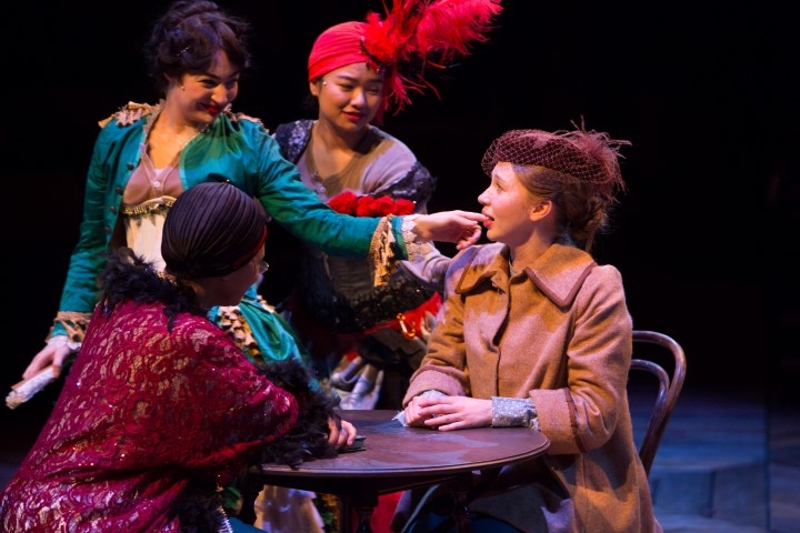 Two women sat at a table. Two women in elaborate bold costumes, including a feather head piece speaking to them stood