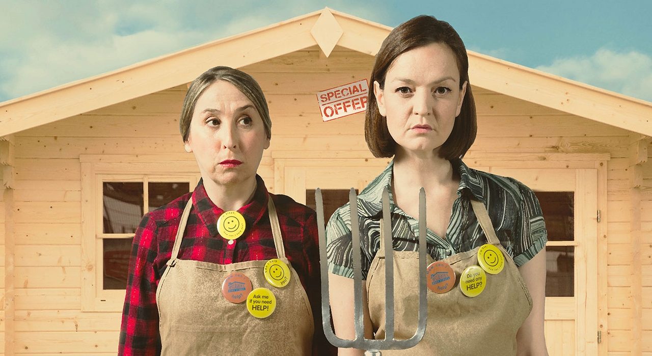 Two women wearing checkered shirts under aprons, One is holding a pitch fork and they are stood infront of a wooden building