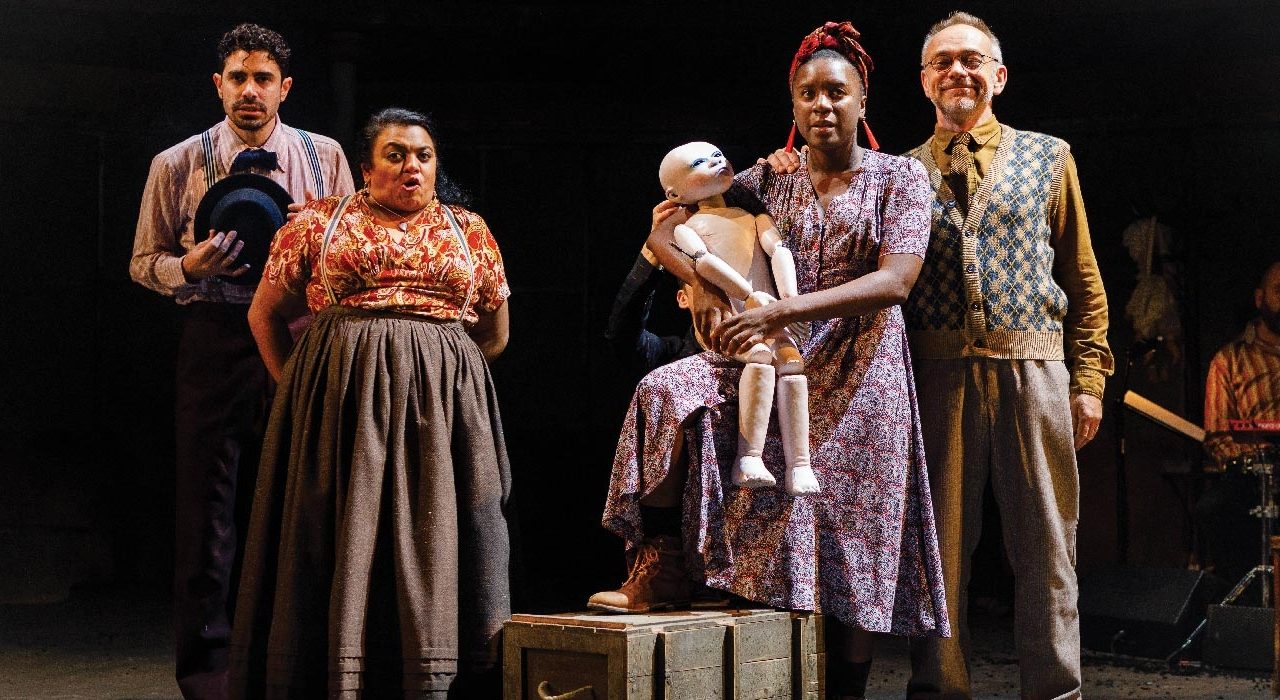 Four people onstage. One is holding a white puppet