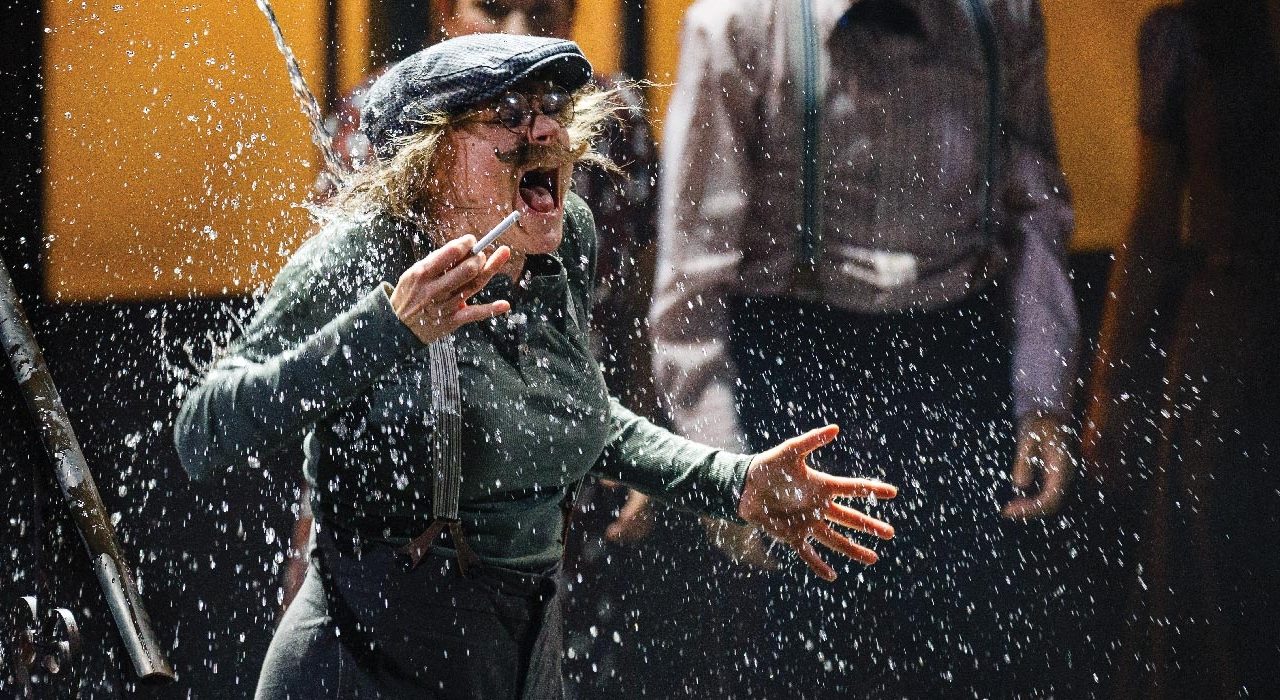 A person in a flat cap, glasses and moustache. being hit by water