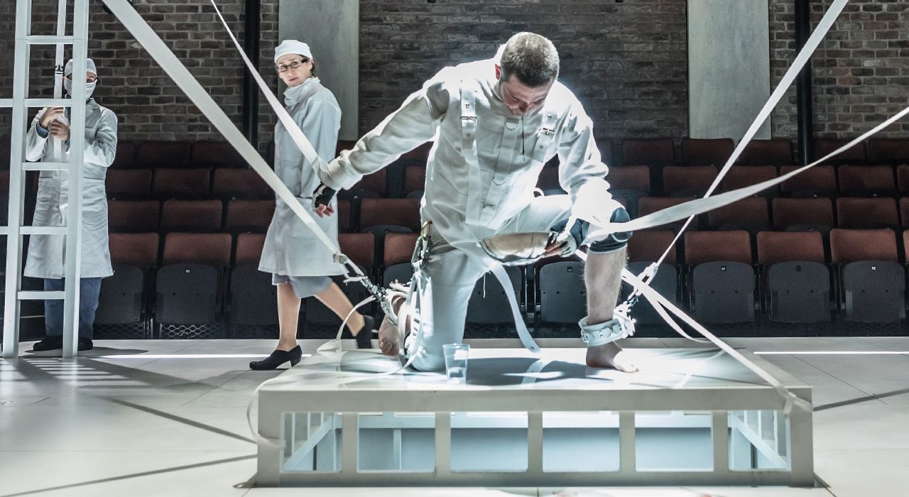 A man kneeling on a platform in an all white almost space suit. There are white strings attached to him.