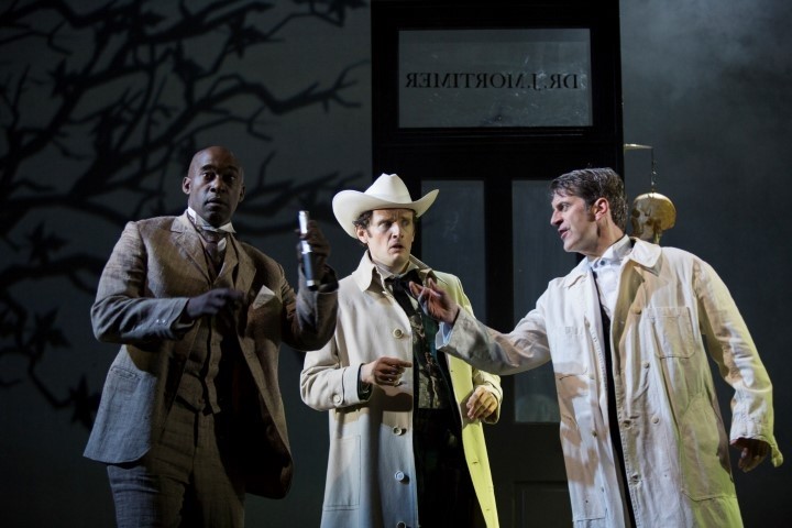 Three men stood in a line. One is wearing a beige three piece suit, one is wearing a trench coat and a cowboy hat, one is wearing a lab coat