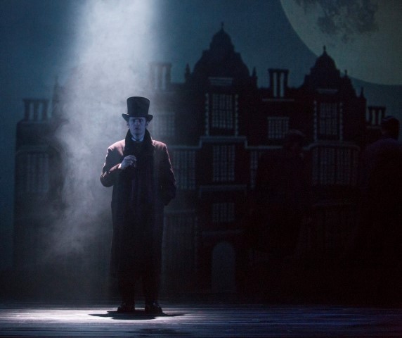 A man stood under a spotlight wearing a top hat and trench coat