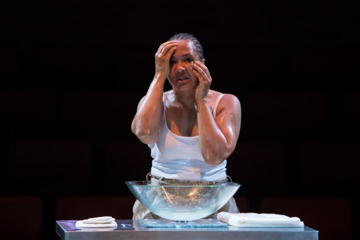 A woman in a white vest washing her face. In front of her is a clear glass bowl and two fluffy towels.