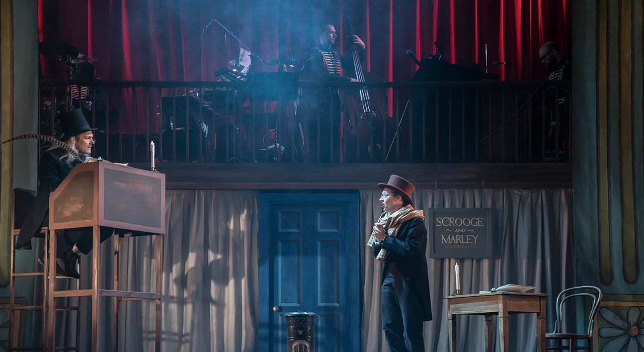 A man sat at a raised desk wearing a suit and top hat holding an oversized quill. He is speaking to another man in a suit, scarf and brown top hat.