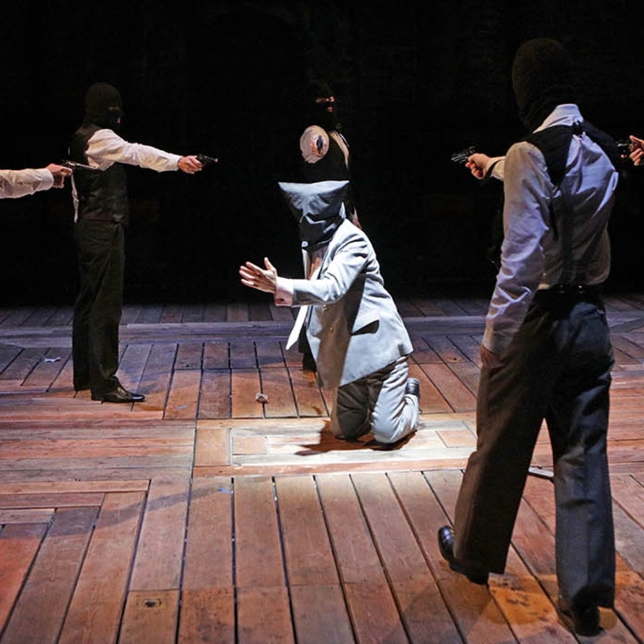 Five people stood in a circle in ski masks, shirts and ties pointing guns at a man kneeling in the centre of the circle on the floor.