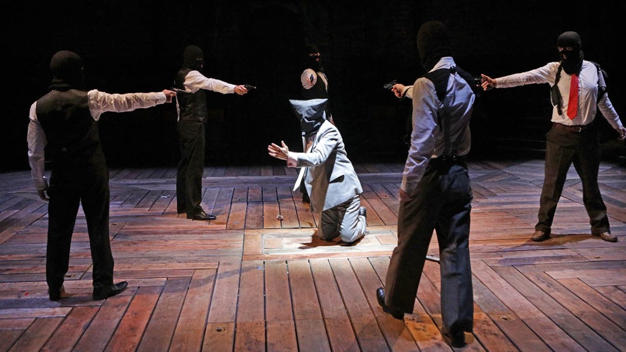 Five people stood in a circle in ski masks, shirts and ties pointing guns at a man kneeling in the centre of the circle on the floor.