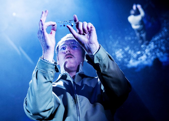 A man with glasses and a moustache is holding a metal wire. There is a mirror behind him reflecting the image,