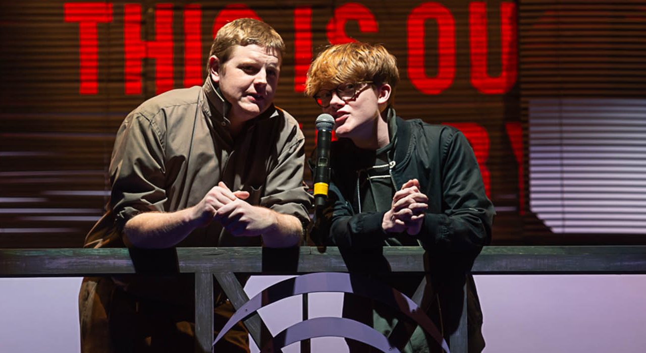 Two people leaning on a bannister speaking into a microphone