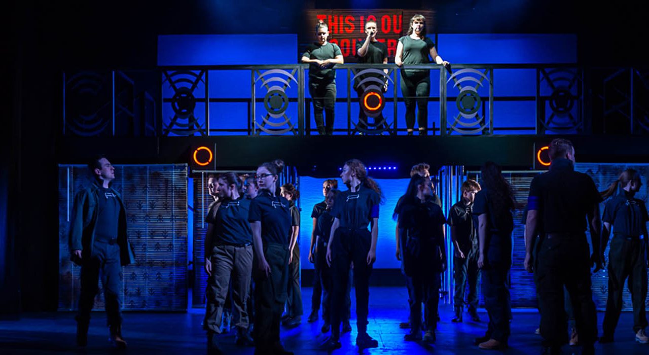 Three people are stood on a bannister overlooking a crowd of the cast.