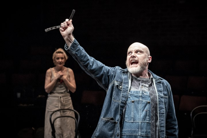 A bald man with a grey beard holding a razor. He is wearing a Blue denim jacket and dungarees.