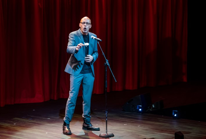 A bald man in a blue suit stood taling in to a microphone