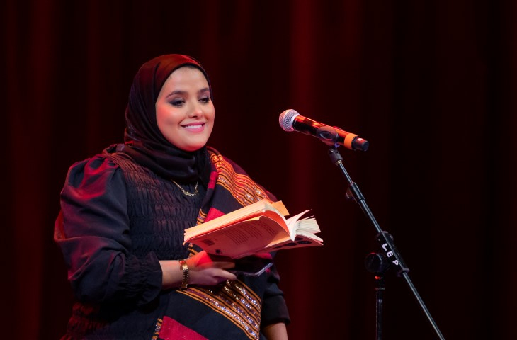 A woman wearing a black head scarf and dress holding a book. She has a red scarf across her body.