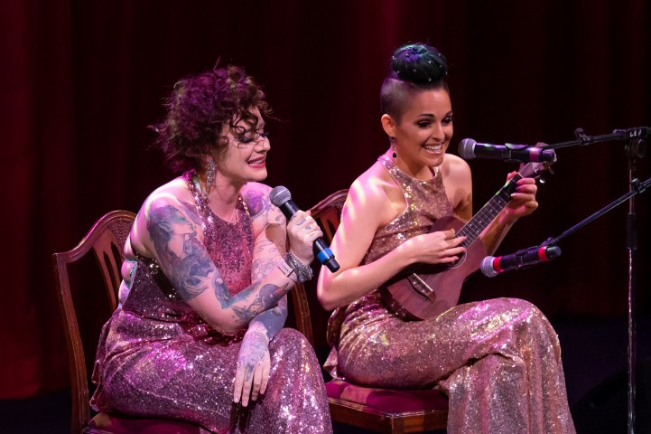 Two woman sat on chairs wearing floor length nude sequin dresses. One is sat with a microphone singing and the other is playing the ukulele and singing.