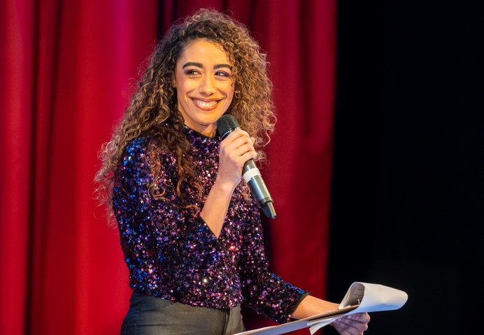 A woman with curly long hair and blonde highlights holding a microphone and clipboard of paper. The woman is wearing a long sleeve purple sequin top and grey trousers.