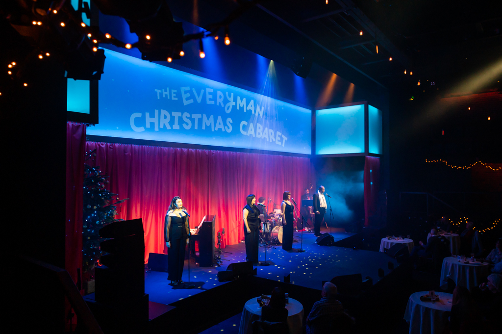 The stage is a red curtain with a light up panel above that says 'The Everyman Christmas Cabaret'