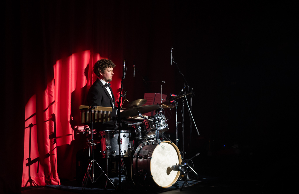 A spotlight of a man playing the drums with a red curtain background