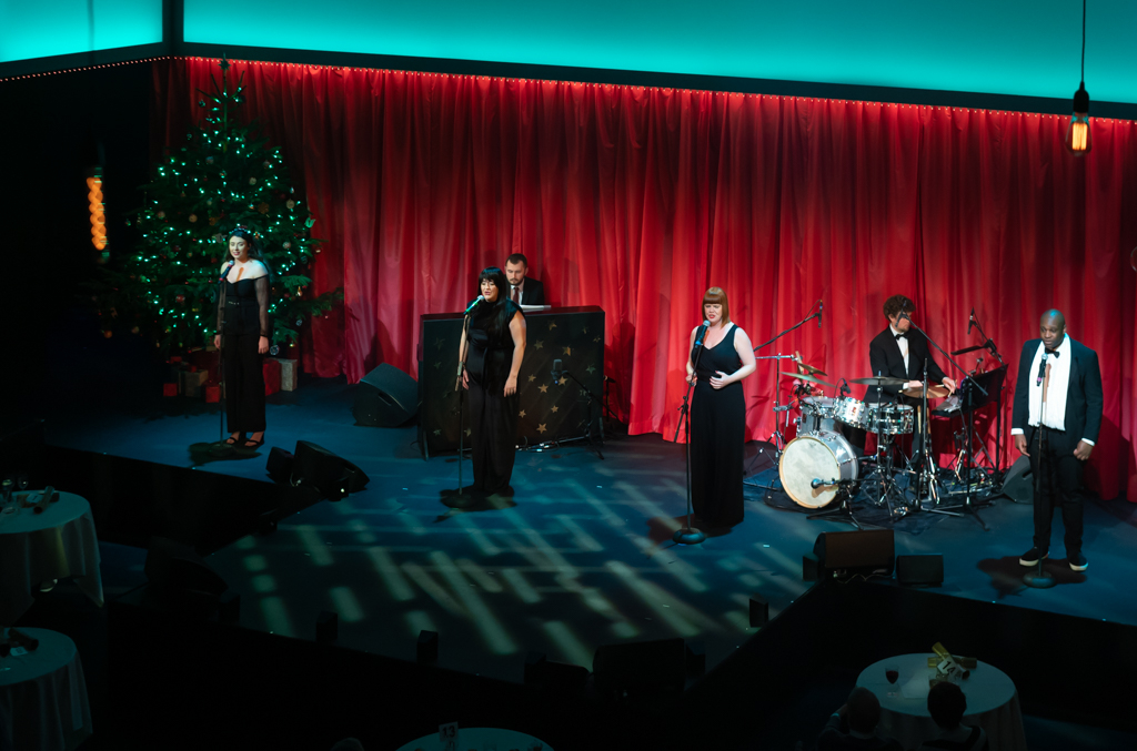 A piano, Christmas tree and drum kit at the back of the stage. At the front of the stage are four singers stood wearing all black.