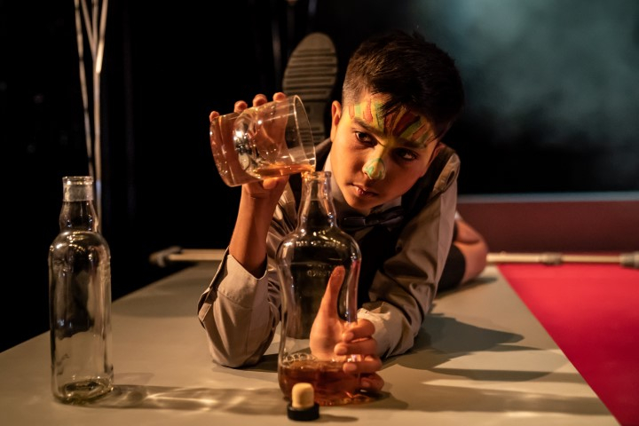 a young boy with face paint on lying on the floor pouring whiskey from the glass back into the whiskey bottle