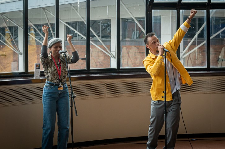 A woman wearing a grey baret, leopard print top and blue jeans, and a man with short brown curly hair, yellow shirt and grey trousers singing in The play house theatre bar.