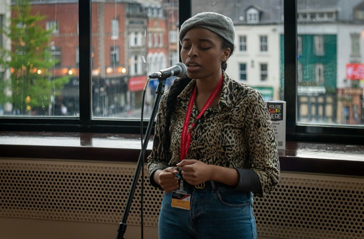 A woman wearing a grey baret, leopard print top and blue jeans singing into a microphone in The Playhouse theatre bar.