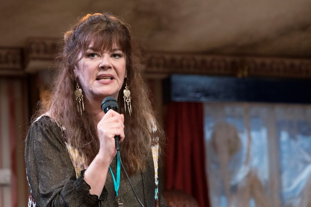 A woman with long brown hair and a full fringe. She is wearing Long gold dangly earrings and has a microphone raised to her mouth